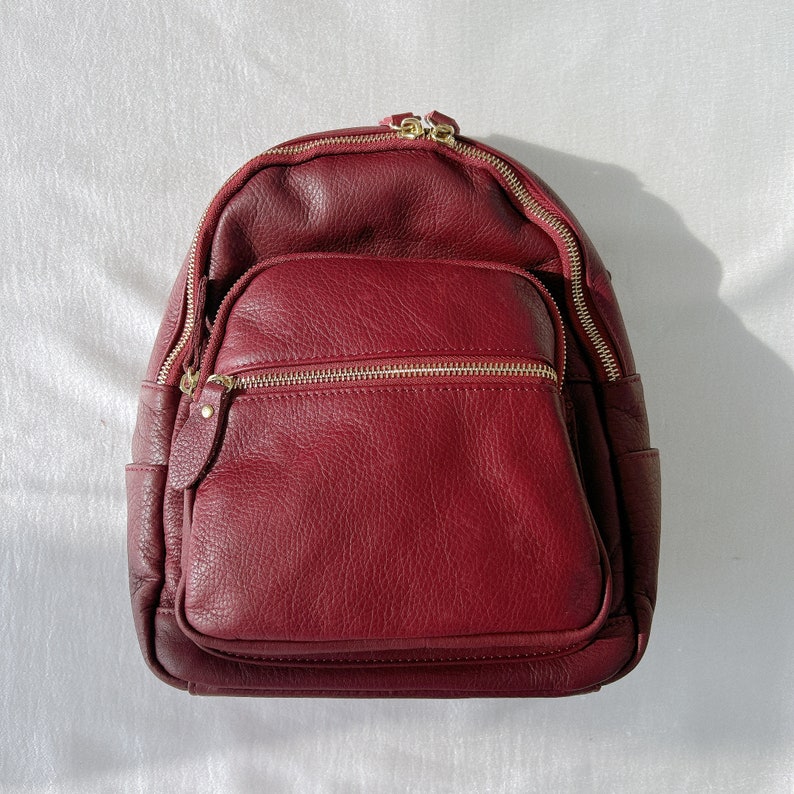 May include: A burgundy leather backpack with gold zippers and a flap closure. The backpack has a textured leather finish and a spacious main compartment.