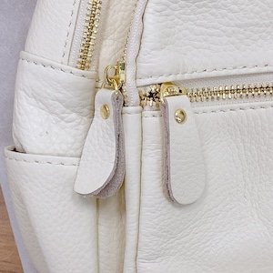 May include: Close-up of a white leather backpack with gold zippers and pulls. The bag has a textured surface and a flap with a zipper closure.