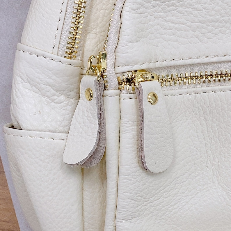 May include: Close-up of a white leather backpack with gold zippers and pulls. The bag has a textured surface and a flap with a zipper closure.