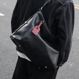 Puede incluir: Bolso de hombro de cuero negro con una etiqueta roja adjunta. El bolso tiene una correa larga y herrajes de color plateado. El bolso se lleva al hombro.