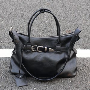 Könnte beinhalten: Eine schwarze Handtasche mit zwei Tragegriffen und einem abnehmbaren Schulterriemen mit Perforationen. Die Vorderseite ist mit einem horizontalen Riemen verziert, der durch zwei silberfarbene Metallschnallen und eine Spitze gesichert ist. Ein kleines rechteckiges Etikett hängt an der Seite.