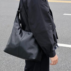 May include: A black hobo-style shoulder bag with a smooth, matte finish and a single wide strap. The bag features a curved top opening, a central vertical seam detail, and a spacious, unstructured body that hangs at the side of the hip.