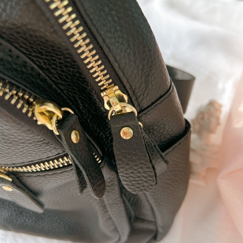 May include: A black leather backpack with gold zippers and pulls. The backpack has a textured leather finish and multiple pockets.