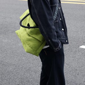 Puede incluir: Un bolso tote de color verde lima con un asa gruesa de cuerda negra trenzada, llevado sobre el hombro. El bolso presenta una estructura de tela suave y texturizada de forma rectangular, combinado con una chaqueta vaquera y pantalón negros.