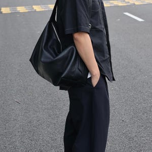 May include: A black leather shoulder bag with a smooth texture and a silver-toned zipper closure. The bag features a slouchy, unstructured silhouette with a wide strap, worn over the shoulder, showcasing a clean design with minimal hardware details.