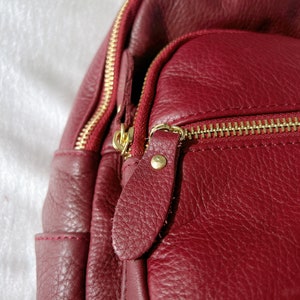May include: Close-up of a burgundy leather backpack with gold zippers and a gold metal zipper pull. The backpack has a textured leather finish.