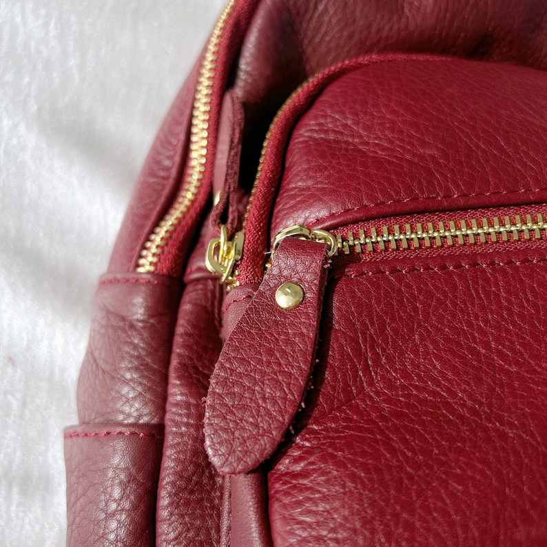 May include: Close-up of a burgundy leather backpack with gold zippers and a gold metal zipper pull. The backpack has a textured leather finish.