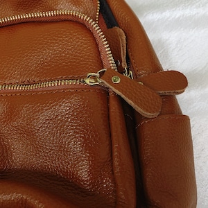 May include: A brown leather backpack with a zipper closure and a leather strap. The backpack has a textured surface and a small pocket on the front.