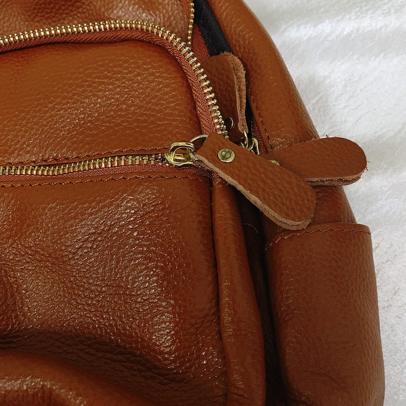 May include: A brown leather backpack with a zipper closure and a leather strap. The backpack has a textured surface and a small pocket on the front.