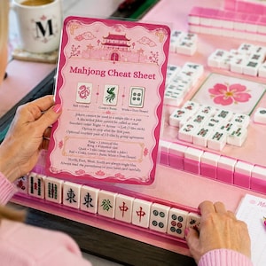 May include: A pink Mahjong cheat sheet with a pagoda illustration, game rules, and tile references. The card is held over a pink Mahjong mat featuring white tiles with green, red, and black symbols, a pink flower tile, and a pink pen on a score sheet.