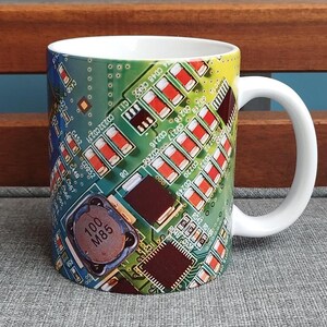 Könnte beinhalten: Eine weiße Keramiktasse mit einem farbenfrohen Design einer Leiterplatte. Das Design zeigt grüne, blaue, gelbe, rote und schwarze Farben. Der Text "100 M85" ist auf der Leiterplatte sichtbar.