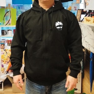 May include: Black zippered hoodie with a white cartoon character on the front. The character has spiky hair and is wearing a headband.