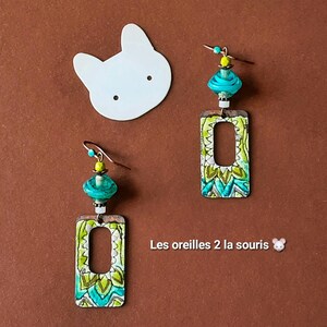 May include: A pair of dangle earrings featuring rectangular metal pendants with a green and teal patterned design. Each earring includes a turquoise glass bead, a small yellow bead, and a metal hook. The text Les oreilles 2 la souris is visible.