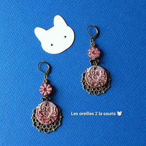 Può includere: Un paio di orecchini pendenti con dischi rotondi testurizzati color rosa polvere, dettagli in filigrana dorata, piccole perline a forma di fiore e chiusure a monachella color bronzo. Il testo Les oreilles 2 la souris è visibile.