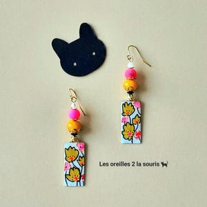 Puede incluir: Pendientes colgantes con diseño floral. Cada pendiente presenta un colgante rectangular con flores amarillas sobre fondo azul claro, acentuado con cuentas rosas y amarillas. El texto "Les oreilles 2 la souris" es visible.