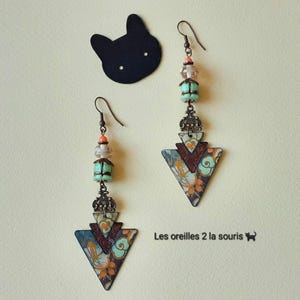 Könnte beinhalten: Ein Paar Ohrhänger mit dreieckigen Metallanhängern mit Blumenmuster in Blau, Orange und Creme. Die Ohrringe enthalten bronzefarbene Filigran-Verbindungsstücke, mintgrüne Perlen, klare facettierte Perlen und kleine orangefarbene Akzente.