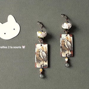 May include: Handmade earrings featuring a heron design. Each earring has a rectangular pendant with a heron illustration, a white flower accent, and a dangling bead. The earrings are set against a gray background with the text "Les oreilles 2 la souris".