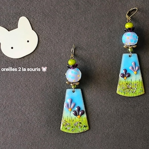 May include: Handmade dangle earrings with a blue, green, and purple floral design. Each earring features a rectangular pendant with a blue sky background, purple flowers, and green grass. The earrings are accented with colourful beads and gold-tone hooks.