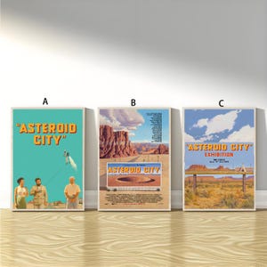 May include: Three framed posters with the title "Asteroid City". The posters feature retro-style illustrations with desert landscapes and characters. The posters are in a row against a white wall, with a wooden floor.