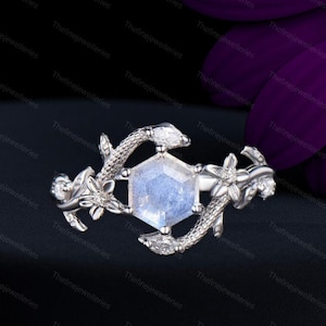 May include: A silver ring featuring a hexagonal, light blue gemstone at its center. The ring's design includes floral and snake-like elements, with small, clear accent stones. The ring is set against a dark background.