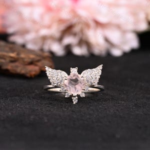 May include: A silver ring featuring a heart-shaped, light pink gemstone at the center, flanked by sparkling, wing-shaped accents. The ring's design includes small, clear stones, creating a delicate and elegant aesthetic. The ring is set against a dark background.
