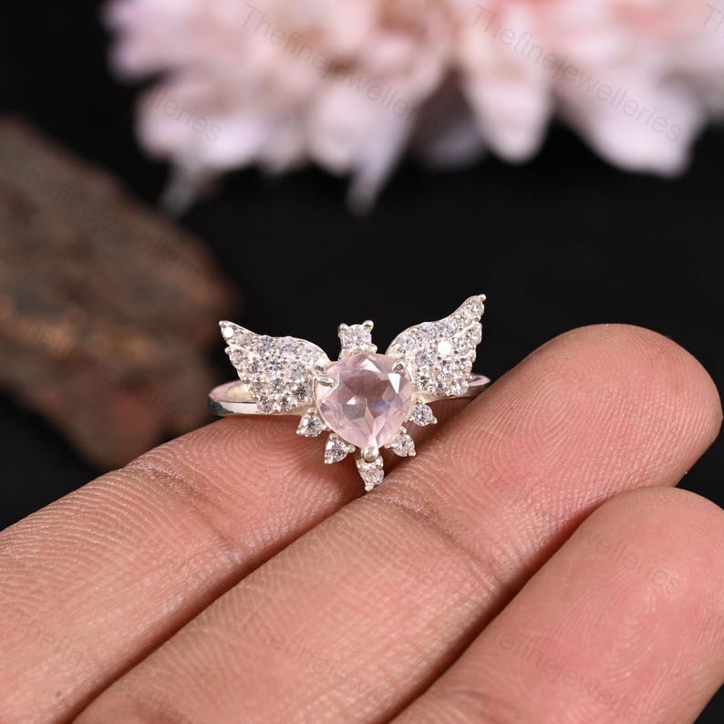 May include: A silver ring featuring a heart-shaped, light pink gemstone, flanked by sparkling, detailed wings. The ring has a halo of smaller, clear gemstones around the heart. The design is elegant and eye-catching, perfect for a special occasion.