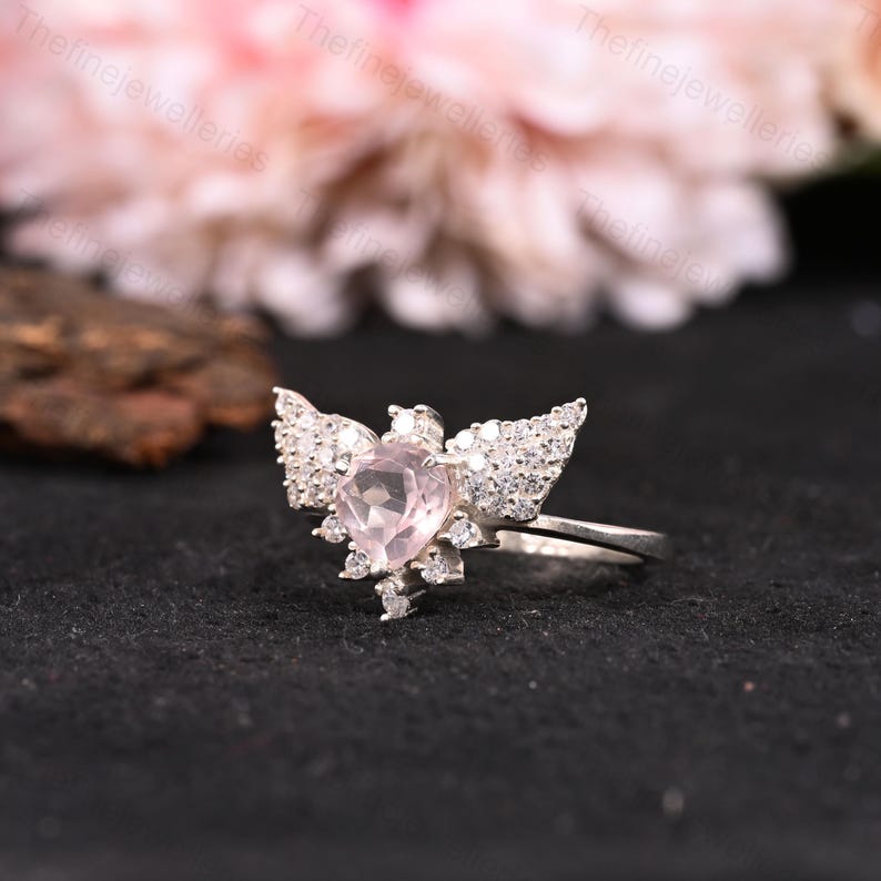 May include: A silver ring featuring a heart-shaped, light pink gemstone at the center, surrounded by smaller, clear gemstones and wing-shaped accents. The ring is set against a dark background, highlighting the sparkle of the stones. The ring is a piece of jewelry.