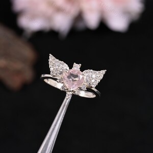 May include: A silver ring featuring a heart-shaped, light pink gemstone at the center, flanked by sparkling, wing-shaped accents. The ring is held by a silver tool against a blurred background.