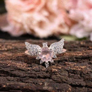 May include: A sterling silver ring featuring a heart-shaped, light pink gemstone, accented with diamond-studded wings. The ring is set against a dark, textured wooden surface, with a blurred floral background.