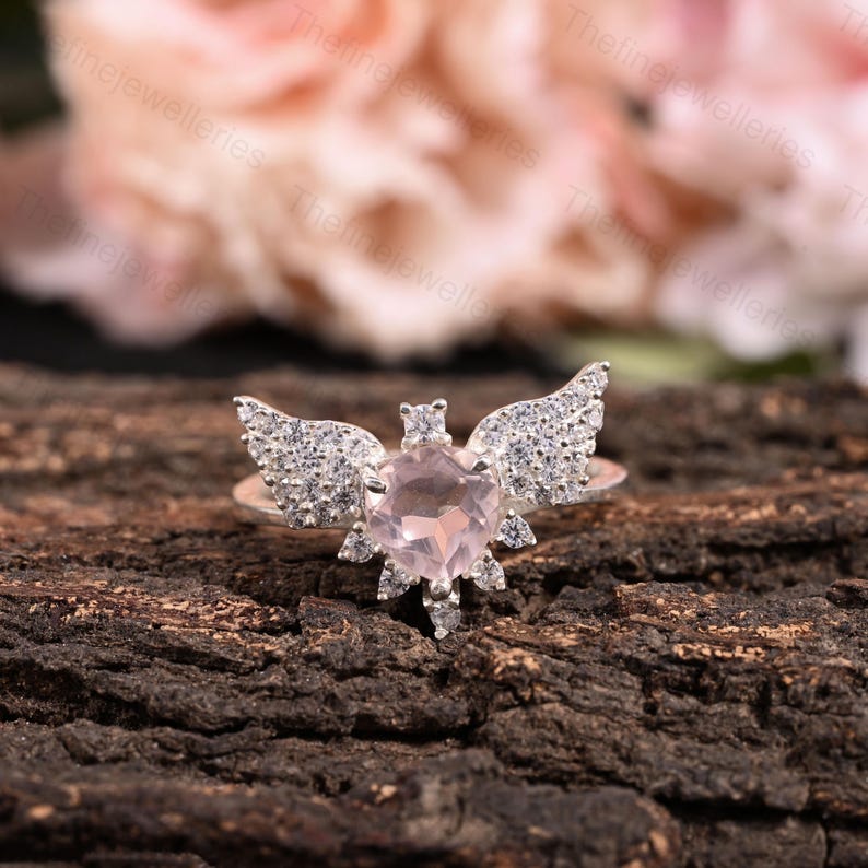 May include: A sterling silver ring featuring a heart-shaped, light pink gemstone, accented with diamond-studded wings. The ring is set against a dark, textured wooden surface, with a blurred floral background.