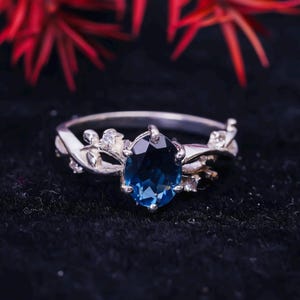 Peut inclure: Une bague en argent avec une pierre précieuse bleue et des accents blancs. La bague a un design délicat en forme de vigne.