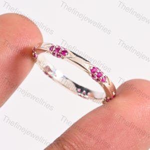 Puede incluir: Un anillo plateado con una fila de pequeñas gemas de color rosa brillante. El anillo tiene un diseño delicado con patrones grabados a lo largo de la banda. Las gemas están espaciadas uniformemente, creando una línea continua de color.
