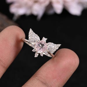 May include: A silver ring featuring a heart-shaped, light pink gemstone at the center, flanked by two wing-shaped clusters of small, clear gemstones. The ring's band is thin and silver.