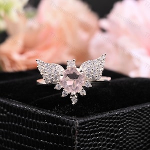 May include: A silver ring featuring a light pink, square-cut gemstone at the center, surrounded by smaller clear stones and wing-shaped accents. The ring is set against a black background, with soft pink and white flowers in the background.