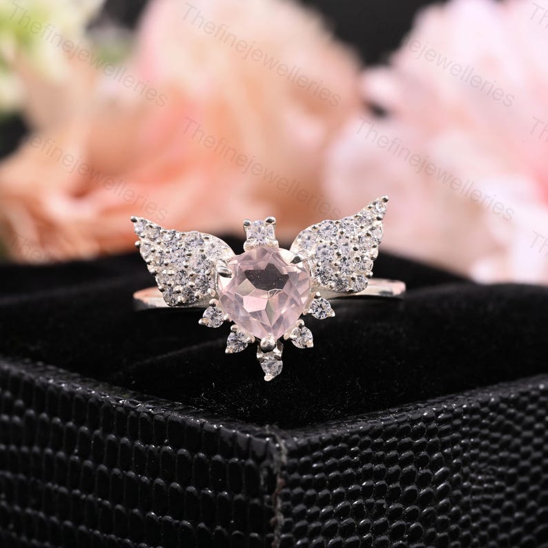 May include: A silver ring featuring a light pink, square-cut gemstone at the center, surrounded by smaller clear stones and wing-shaped accents. The ring is set against a black background, with soft pink and white flowers in the background.