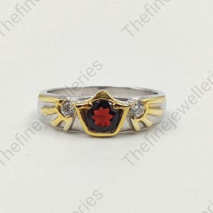 May include: A gold and silver ring with a red gemstone and two smaller clear gemstones. The ring has a decorative design with wings on either side of the center stone.