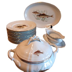 Può includere: Un set di stoviglie in porcellana bianca con un design a forma di pesce. Il set comprende una zuppiera con coperchio, una salsiera e una pila di piatti.