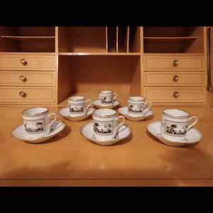 Puede incluir: Juego de seis tazas y platillos de espresso de porcelana blanca. Cada taza y platillo presenta un diseño de paisaje en blanco y negro y ribete dorado. Las tazas tienen asas pequeñas y están sobre una superficie de madera.
