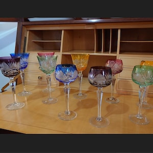 Puede incluir: Una colección de copas de vino de cristal de colores con intrincados diseños tallados. Las copas presentan varios tonos, incluyendo morado, azul, verde, amarillo y rojo. Cada copa tiene un tallo largo y una base redonda, sobre una superficie de madera.