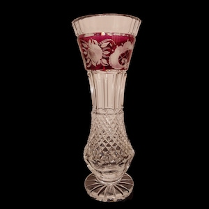 Può includere: Un vaso di vetro trasparente con un designo in cristallo tagliato. Il vaso ha un'ampia apertura e una base stretta. La parte superiore del vaso è decorata con un motivo floreale rosso e bianco.