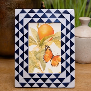 May include: A rectangular picture frame with a geometric pattern of white and navy blue triangles. The frame holds a print of an orange and a butterfly. The frame is on a wooden surface.