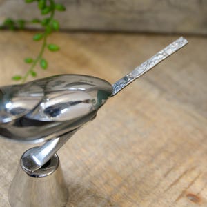 May include: A polished silver bird figurine with a detailed wing and tail design. The bird is mounted on a conical base, and the background is a wooden surface. The figurine is a decorative item.