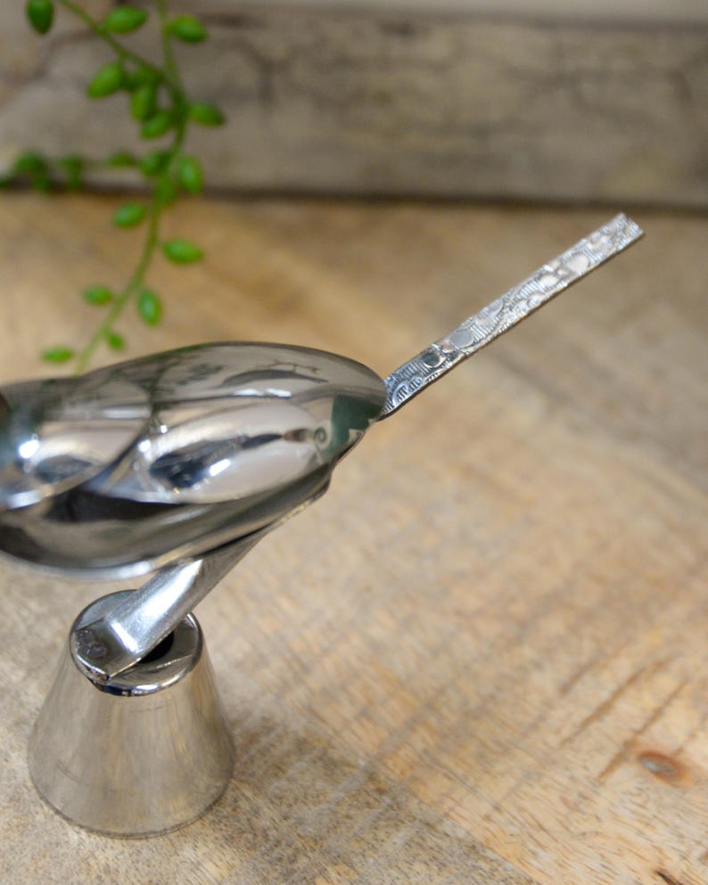 May include: A polished silver bird figurine with a detailed wing and tail design. The bird is mounted on a conical base, and the background is a wooden surface. The figurine is a decorative item.