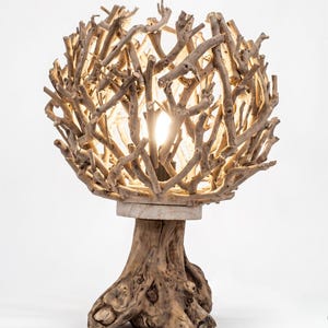 May include: A table lamp featuring a spherical shade constructed from numerous light brown driftwood branches. The base is a solid, textured piece of natural wood stump. A warm light glows from within the centre of the branch structure.