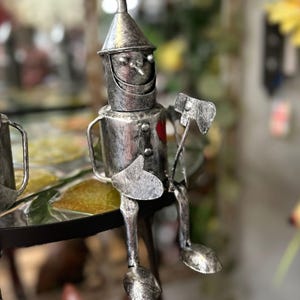 May include: A silver-colored metal figurine of the Tin Man from The Wizard of Oz. The figurine has a conical hat, a red heart, and is holding an axe. It is sitting with its legs dangling.