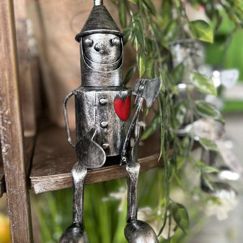 May include: A metallic silver figurine of the Tin Man from The Wizard of Oz, with a red heart and an axe. The figurine is seated and has a conical hat. The background includes greenery and a wooden shelf.