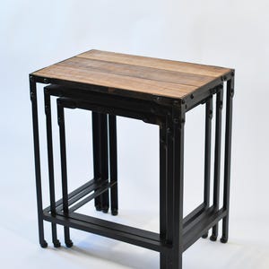 May include: A set of three nesting tables. Each table features a rectangular, light brown wooden top and a black metal frame. The frames have straight legs and horizontal supports, creating a modern, industrial look. The largest table is approximately 61 cm wide.