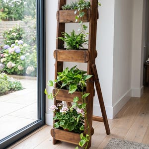 May include: A tall, dark brown wooden ladder-style plant stand featuring four horizontal shelves. Each shelf holds a variety of potted green plants, including trailing ivy, ferns, pothos, and pink flowering geraniums, arranged vertically.