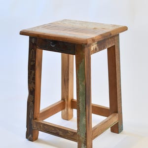 May include: A square, rustic wooden stool with a small drawer. The stool has a natural wood finish with visible grain and a mix of colours, including brown, green, and blue. The legs are sturdy and angled, providing stability.