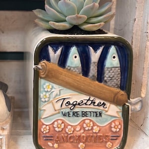 May include: A ceramic planter shaped like a rectangular tin of anchovies with a succulent plant inside. The front features a relief design of two fish, a brown key, and the text "Together WE'RE BETTER ANCHOVIES" surrounded by small white flowers.